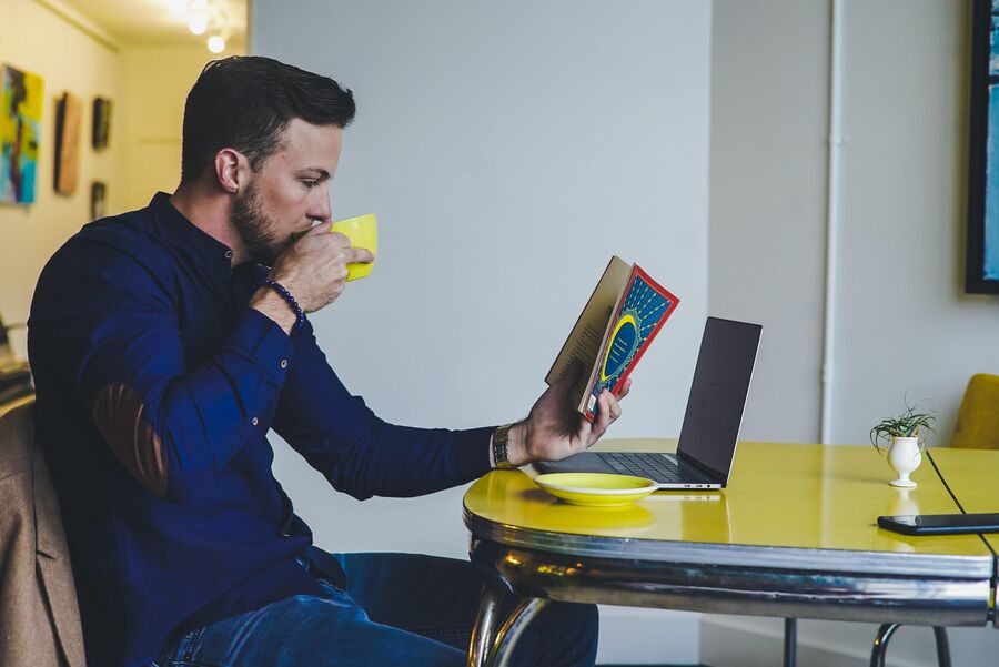 En mand drikke kaffe og læser en bog ved sin computer. Foto: Chris Benson for Unsplash.