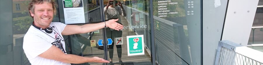 Thomas står og peger på turistinformationsskiltet på døren til Næstved Bibliotek